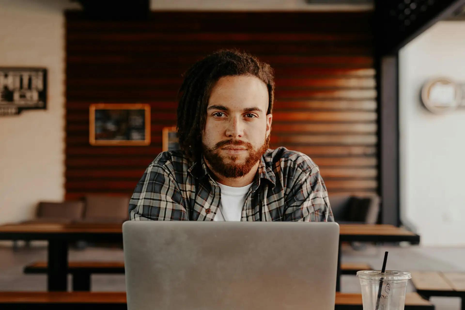 Person with beard and plaid shirt working on laptop in a cozy café with wooden interior