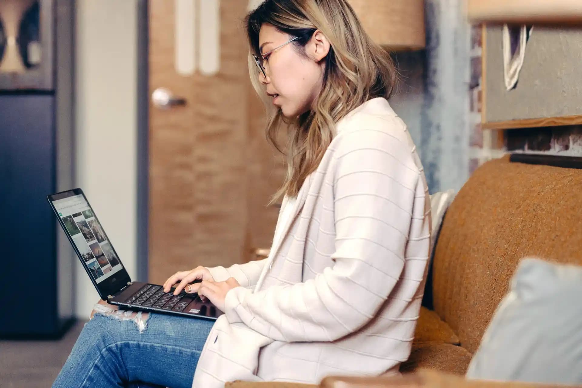 Person mit Brille in einem hellen Pullover die auf einer Couch sitzend Stellenanzeigen auf einem Laptop durchsucht