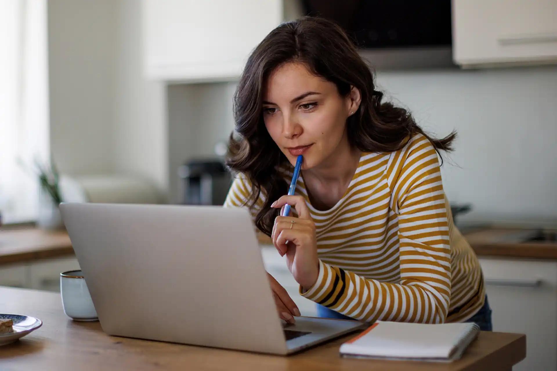 Young woman thoughtfully working on a laptop from home with a pen to her lips
